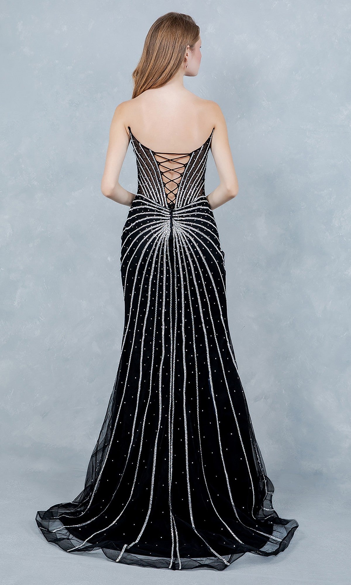 A woman wearing the Cinderella Couture 8576J long prom dress with silver embellishments and a lace-up corset bodice stands facing away against a light gray background.